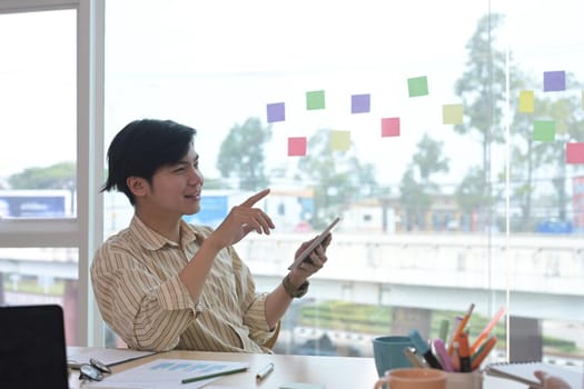 Handsome asian businessman sitting at creative workplace and using digital tablet.