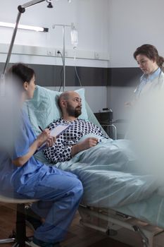 Nurse sitting at patient bedside writing prescription medication for pain relief using clipboard. Middle aged man recovering after surgical intervention talks about treatment with doctor.