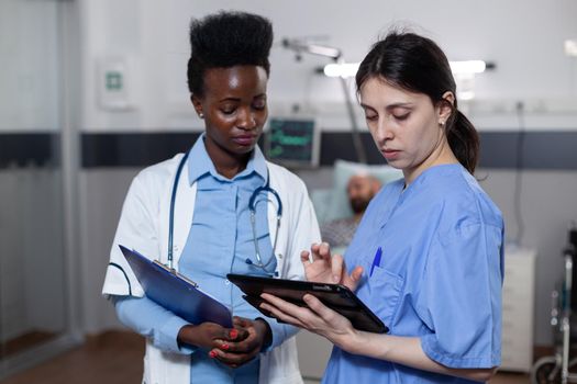 Imaging operator showing scan results on digital tablet to medic holding clipboard with lab results in modern hospital ward. Health care specialists using modern technology for clinical diagnosis.