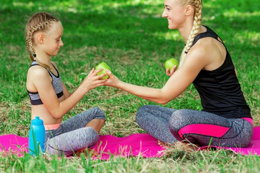 Mother is giving to daughter a green apple sitting on the roll mat in park.