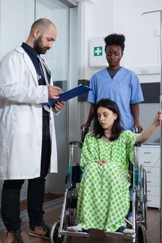 Nurse moving patient sitting in wheelchair holding iv pole to treatment room while doctor gives consult looking at clipboard with lab results. Woman with illness being checked before surgery.