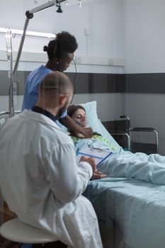 Doctor during morning rounds checking clipboard with patient records while nurse is putting oxygen mask on unconscious woman. Caregiver attending recovering female in modern hospital ward.