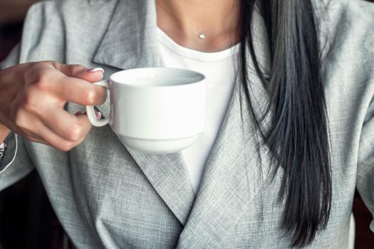 Young businesswoman is holding a white cup of hot drink wearing jacket, close up.