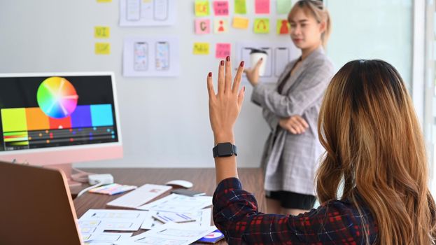 Rear view of creative woman raised hand up for asking question during designer team meeting.