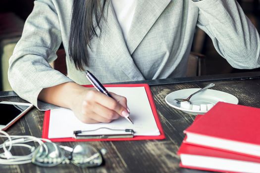 Business woman is writing in a sheet of paper wearing a jacket sitting in cafe.