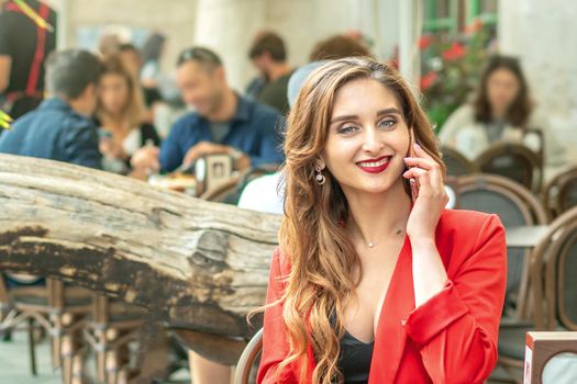 Young cute girl is talking by mobile phone sitting alone in coffee shop outdoors.