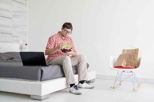 Online shopping concept. Young man sitting on the bed and shopping online using tablet looking at the credit card, copy space