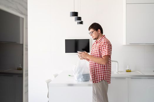 Delivery food, products to home. Shopping and healthy food concept. Young man in red plaid shirt holding a disposable plastic bag with food delivery at the modern kitchen