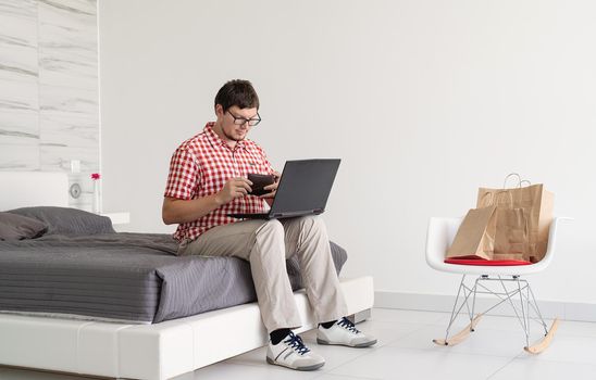 Online shopping concept. Young man sitting on the bed and shopping online using tablet looking at the credit card, copy space