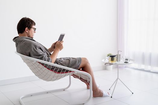 Young caucasian man in gray bathrobes sitting in the living room relaxing and drinking green tea, using tablet