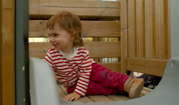 Little girl sitting on a top of a wooden slide and smiling. Baby on the playground in the park.