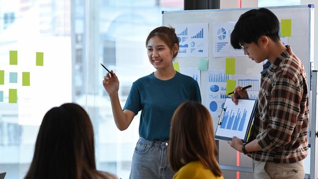 Young creative female standing near flip chart with her colleague and giving presenting new marketing strategy in office.