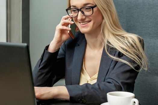 Beautiful young smiling woman with glasses is talking on the cell phone working on a laptop in home office.