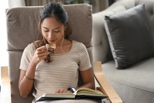 Satisfied asian woman drinking coffee and reading book on armchair at home.