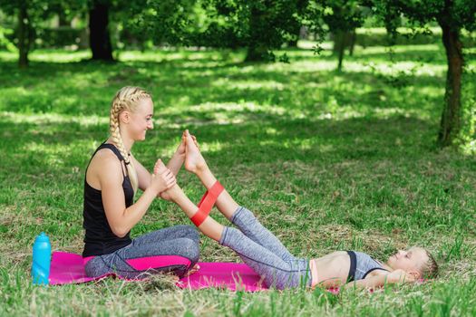 Mother is training daughter's legs by fitness gum on mat in the park.