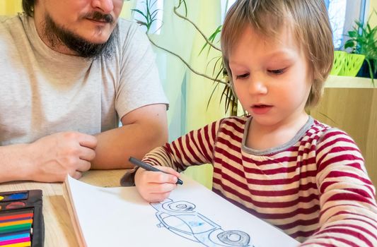 Bearded man in grey t-shirt and cute three - year - old boy drawing pictures with colored wax crayons in a cozy yellow room while spending time at home together. High quality photo
