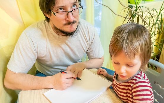 Bearded man in grey t-shirt and cute three - year - old boy drawing pictures with colored wax crayons in a cozy yellow room while spending time at home together. High quality photo