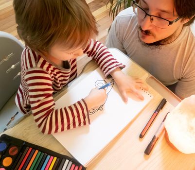 Bearded man in grey t-shirt and cute three - year - old boy drawing pictures with colored wax crayons in a cozy yellow room while spending time at home together. High quality photo