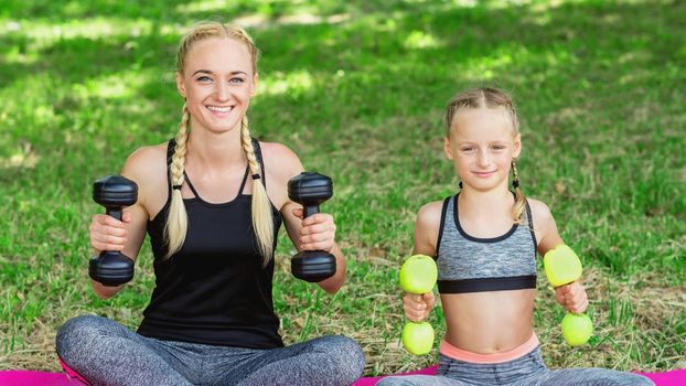 Young mother with her little daughter are training with apple dumbbells in the park.