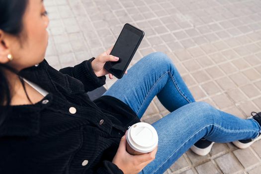 unrecognizable asian girl typing on a phone sitting outdoors on a concrete floor