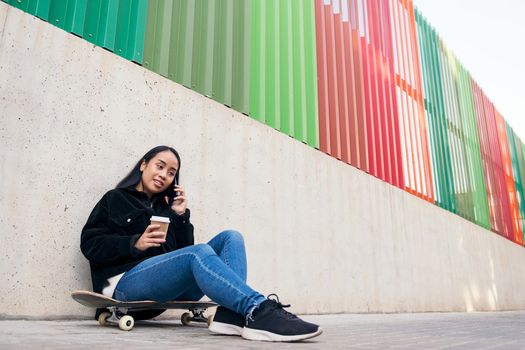 asian teenage girl talking by phone outdoors sitting on her skateboard leaning against a concrete wall
