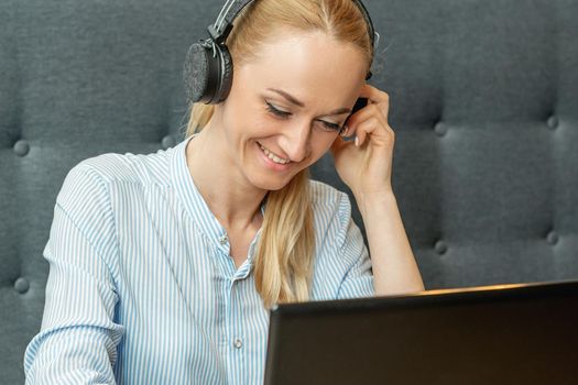 Happy young woman wearing headphones looking on laptop screen during online training with video conferencing at home office.