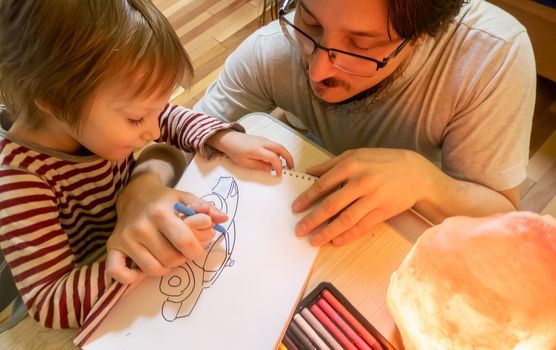 Bearded man in grey t-shirt and cute three - year - old boy drawing pictures with colored wax crayons in a cozy yellow room while spending time at home together. High quality photo