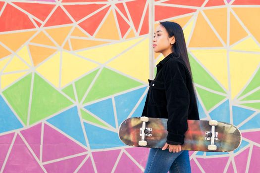 young asian woman walks with a skateboard in her hand in front of a colorful wall