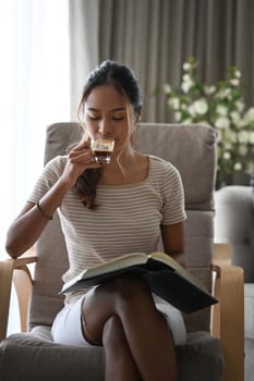 Casual young woman drinking coffee and reading book while spending leisure time at home.