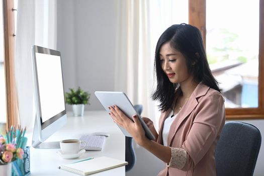 Concentrated young businesswoman sitting at modern workplace and using digital tablet.