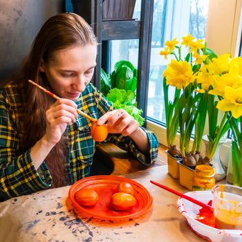 woman draws paper rabbit ears. Preparation for Easter: a young woman paints paper rabbit ears with paints in the kitchen among fresh greenery, flowers and painted eggs