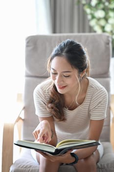 Millennial asian woman sitting in comfortable armchair and reading her favorite novel book.