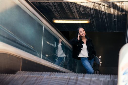 young asian woman rides escalator at subway station with skateboard in hand while talking on phone