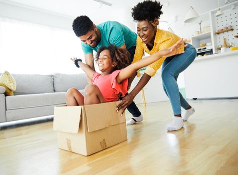 Mother and father pushing their daughter sitting in a cardboard box, having fun at home