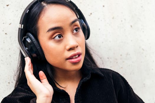 young asian woman enjoys listening to music with headphones outdoors leaning against a concrete wall