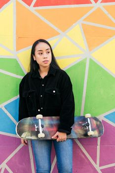young asian woman with her skateboard leaning against a colorful wall