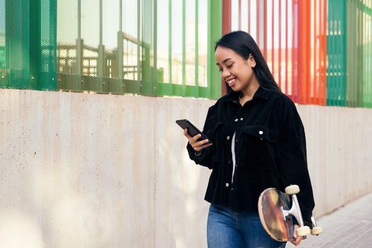 smiling young asian girl typing on the mobile phone while walking down the street with her skateboard in her hand