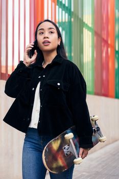 vertical photo of a young asian girl talking on the phone while walking down the street with her skateboard in her hand