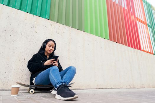 smiling asian girl with headphones listening music from phone sitting outdoors on her skateboard leaning against a concrete wall