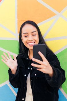 young asian woman greets by making a video call with her cell phone next to a colorful wall