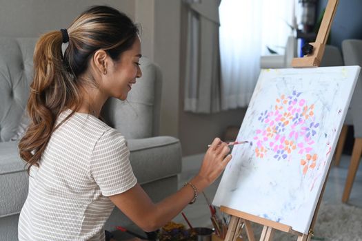 Smiling young asian woman sitting in front of easel and painting picture with water color on canvas.