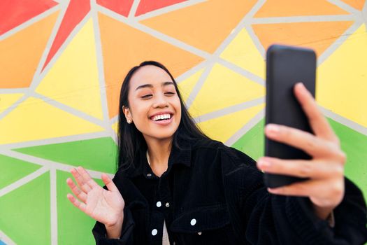 happy young asian woman says hello with the hand making a video call with her mobile phone next to a colorful wall