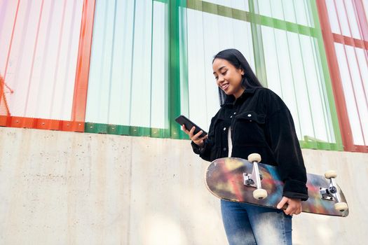smiling young asian girl typing on the mobile phone standing outdoors with her skateboard in her hand
