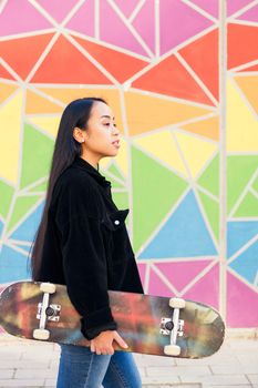 young asian woman walking with a skateboard in her hand in front of a colorful wall