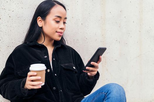 young asian woman with a coffee typing on a phone sitting outdoors leaning against a concrete wall