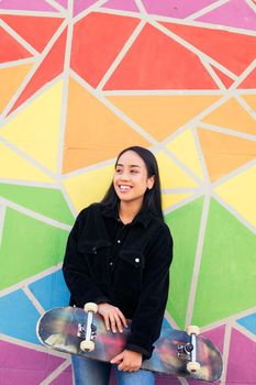 vertical photo of a smiling asian woman with her skateboard leaning against a colorful wall