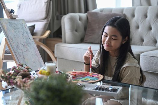 Pretty young asian girl in casual clothes sitting in living room and painting picture with watercolor.