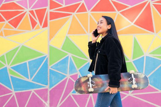 smiling young asian woman talking by phone while walks with a skateboard in her hand in front of a colorful wall
