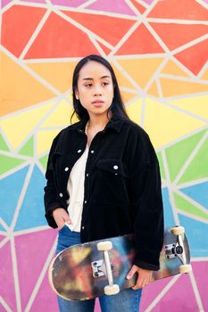 young asian woman with a skateboard in her hand standing in front of a colorful wall