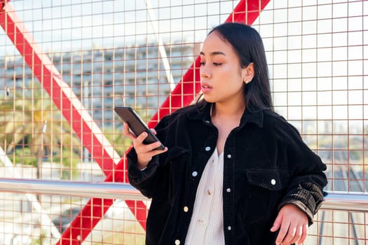 young asian woman outdoors consulting her cell phone leaning against a red fence